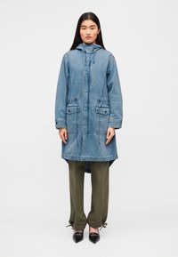 Μακρύ τζιν μπουφάν με κουκούλα με κορδόνι, κούμπωμα με κουμπιά, δύο τσέπες μπροστά, καμπυλωτό τελείωμα και πράσινο παντελόνι συνδυασμένο με μαύρα μυτερά παπούτσια.