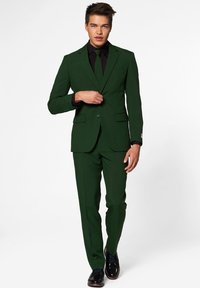 Abito verde caratterizzato da una giacca attillata e pantaloni stretto, abbinato a una camicia nera e una cravatta coordinata. Scarpe nere lucide completano il look.