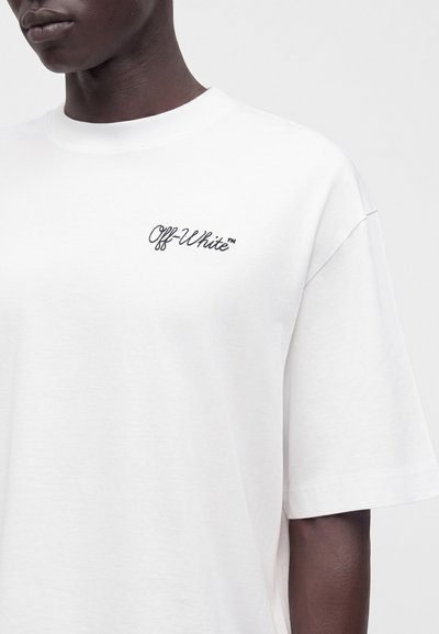 Personne portant un t-shirt blanc à manches courtes avec un petit logo texte noir "Off-White" sur la poitrine gauche, sur un fond uni.