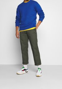 Niebieski sweter dzianinowy z okrągłym dekoltem nałożony na żółtą koszulę, zestawiony z oliwkowymi spodniami i białymi trampkami z kolorowymi akcentami.