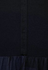 Marinblått tyg med en slät stickad textur, kombinerat med en rynkad genomskinlig nederdel. En märkt satinsticka löper vertikalt längs sömmen på sidan.