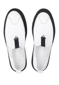 Weiße Slip-On-Schuhe mit einem strukturierten Lederobermaterial, schwarzer Gummisohle und einem dreieckigen metallischen Akzent vorne.