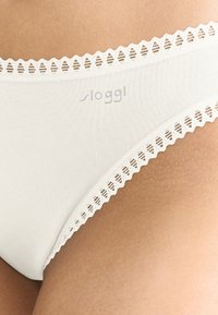 Valkoiset puuvillaiset bikinihousut, joissa on aaltoileva pitsireunus, sileä pinta ja hienovarainen logo vyötärönauhassa.