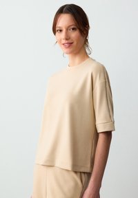 Beżowa bluzka z krótkim rękawem i okrągłym dekoltem, z podwiniętymi mankietami oraz subtelnymi detalami szycia, wykonana z miękkiego, teksturowanego materiału.