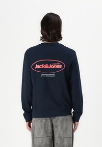 Marineblaue Sweatshirt mit gerippten Bündchen und Saum, auf dem Rücken mit einem großen roten Logo "Jack & Jones Club1990" bedruckt. Aus Baumwollmaterial.