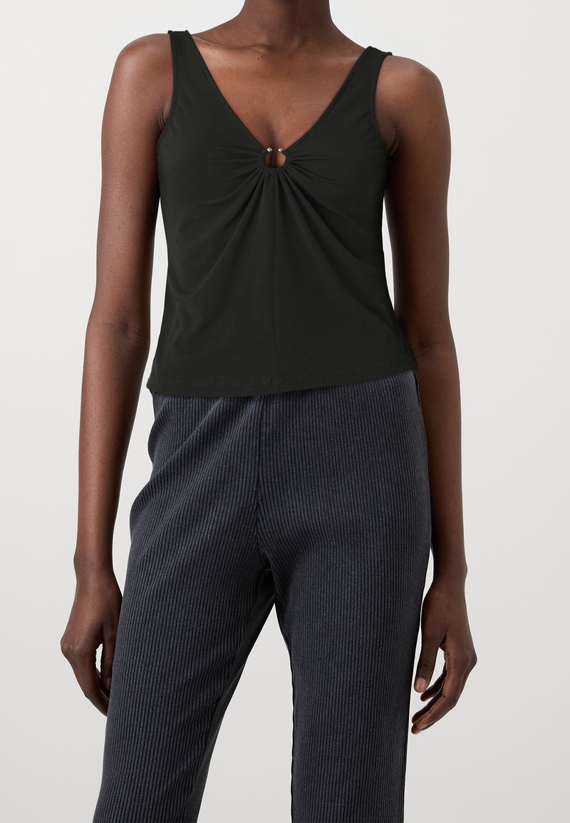 Débardeur noir avec un décolleté en V et un détail central torsadé comportant une petite accent métallique. Associé à un pantalon large en maille côtelée foncé.