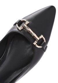 Scarpa in pelle nera a punta con accentuazione in metallo dorato e dettaglio a cintura sulla parte frontale. Texture liscia, design elegante e stile minimalista.