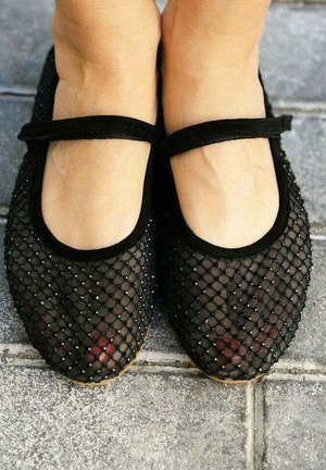 Pies con zapatos Mary Jane de malla negra con tiras, parados sobre un pavimento de baldosas grises.