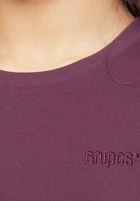 Bordeaux T-skjorte med ribbet tekstur, rund hals og brodert "snipes"-logo på venstre nederste side. Myk stoff, tettsittende design.