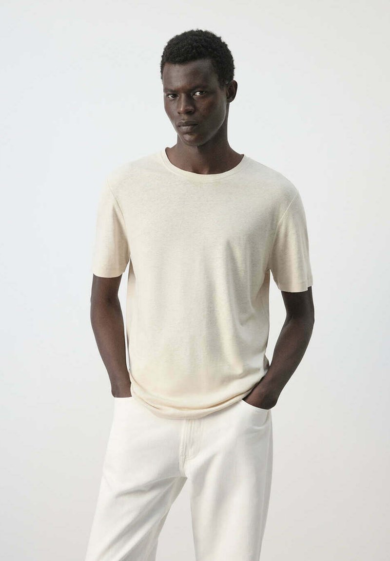 Jeune homme à la peau foncée portant une chemise beige à manches courtes et un pantalon blanc, debout les mains dans les poches sur un fond clair.