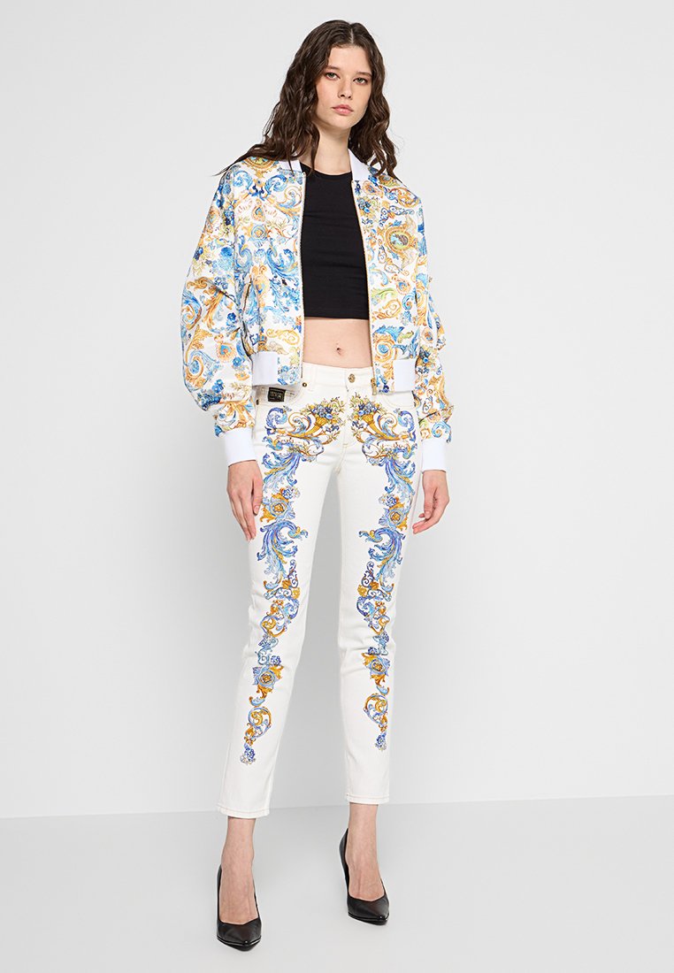 Weiße bestickte Hosen mit blauen und goldenen ornativen Designs, kombiniert mit einer gemusterten Bomberjacke in Blau, Orange und Cremefarben.