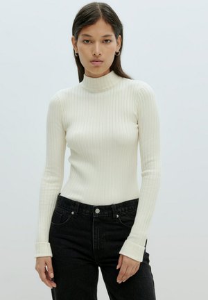 Weißer gerippter Pullover mit Rollkragen, langen Ärmeln, taillierter Passform und umgeschlagenen Bündchen, kombiniert mit hochgeschnittenen schwarzen Jeans.
