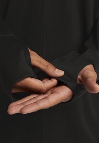 Czarny longsleeve z długim rękawem i otworami na kciuki, wykonany z teksturowanej tkaniny i dopasowany do sylwetki. Zbliżenie pokazuje ręce ułożone na mankiecie.