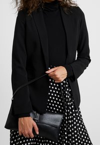 Žena ve černém blejzru a roláku, držící černou texturovanou kabelku přes černou sukni s bílými puntíky.