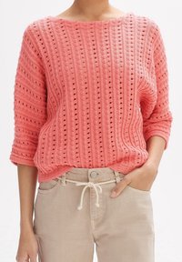 Koraalroze gebreide trui met een brede halslijn, getextureerde horizontale en verticale strepen, en gaatjespatronen. Gecombineerd met lichtbeige broek.