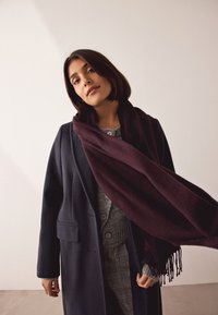 Nāvijas mētelis ar priekšējo kabatu, pārslāņots virs pelēka adīta džempera, pievienota tumši sarkana šalle, kas ir brīvi uzlikta ar bārkstīm.