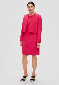 Fuchsia roze jurk met een cropped jasje, beiden van gladde stof. Zwarte hakken mules met gekruiste banden maken de look af.