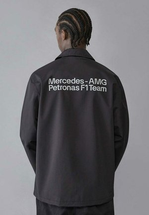 Czarna kurtka z kołnierzykowym designem. Na plecach białe napisy: "Mercedes-AMG Petronas F1 Team". Gładka tekstura materiału.