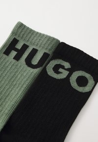 Deux paires de chaussettes : l'une en vert texturé avec "HUGO" en lettres noires en gras, l'autre en noir uni avec "HUGO" en police hexagonale verte.