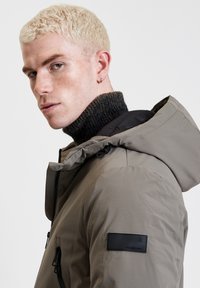Giacca con cappuccio in materiale grigio chiaro con colletto scuro testurizzato, caratterizzata da una patch con logo sul braccio sinistro e tasche laterali con zip.