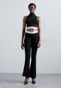Black knitted two-piece set featuring a sleeveless turtleneck top and flared pants, with a logo-embellished waistband. Heels add length.