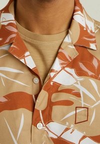 Camicia a maniche corte color beige con stampa di foglie marroni. Presenta un colletto a punta, chiusura con bottoni e un dettaglio ricamato a forma di quadrato sul petto.