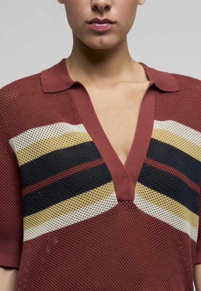 Femme portant un polo en maille filet couleur rouille avec un col en V profond et des rayures horizontales crème, moutarde et bleu marine sur la poitrine.