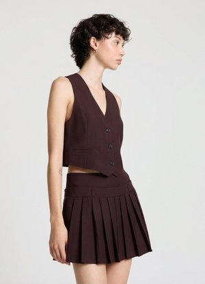 Jeune femme aux cheveux courts et bouclés porte un gilet sans manches boutonné marron foncé et une mini-jupe plissée assortie, debout de profil.