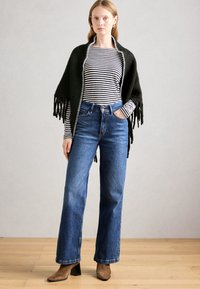 Frau steht auf einem Holzfußboden und trägt einen schwarzen Fransen-Schal, ein schwarz-weiß gestreiftes Langarm-Oberteil, blaue Jeans und braune Absatzstiefel.