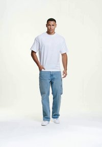 Jovem vestido com uma t-shirt branca, calças cargo de cor azul claro, e sapatilhas brancas, em pé com uma mão no bolso contra um fundo claro e simples.