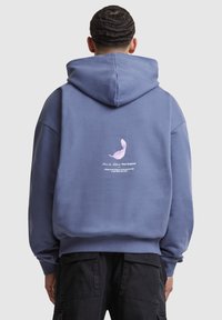 Oversized blue hoodie with a kangaroo pocket, featuring a pink feather graphic and white text on the back. Soft cotton material.
