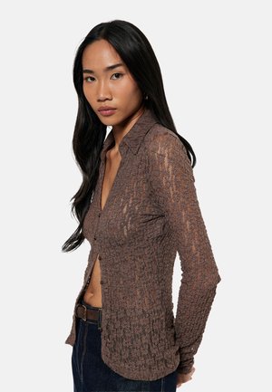 Jonge vrouw met lang zwart haar, gekleed in een doorschijnend bruin gestructureerd overhemd met knopen en donkere jeans, staand tegen een effen lichte achtergrond.