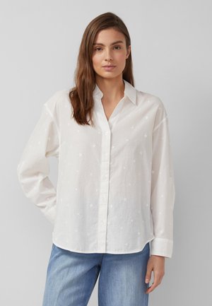 Vrouw draagt een witte blouse met knopen en subtiele sterpatronen, gecombineerd met een blauwe spijkerbroek, staande tegen een effen grijze achtergrond.