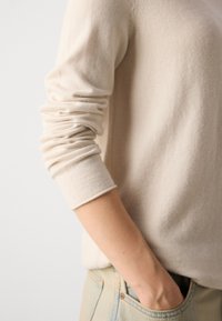 Beige genser laget av mykt stoff, med lange ermer med rullemansjetter, kombinert med lyseblå denimjeans. Glatt tekstur.
