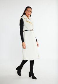 Witte nepbont lange vest met een sjawlkragen en een ceintuur in de taille. Gekocht boven een zwarte top en gecombineerd met zwarte enkellaarzen.