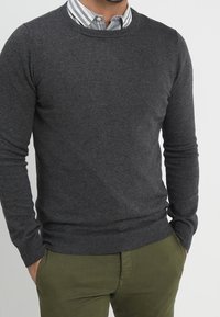 Dunkelgrauer Pullover mit Rundhalsausschnitt aus weichem Strickstoff, mit gerippten Bündchen und Saum, getragen über einem hellblau-weiß gestreiften Hemd.