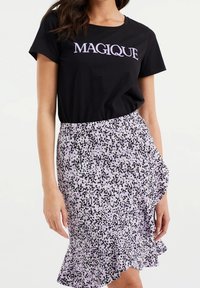 Černé tričko s textem "MAGIQUE", spárované s lilacovou sukní se vzorem černých skvrn a volánkovým lemem. Neformální a vrstvený design.