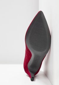Zapato de tacón alto de ante rojo con puntera afilada, suela negra texturizada y detalles mínimos de marca en la parte inferior.
