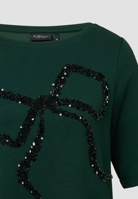 Groene T-shirt van zachte stof met een decoratief zwart pailletten strikontwerp aan de voorkant, korte mouwen en een ronde halslijn.
