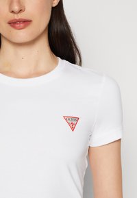 T-shirt in cotone bianco con un piccolo logo triangolare rosso che presenta il testo "GUESS" e un punto interrogativo. Maniche corte e scollo rotondo.