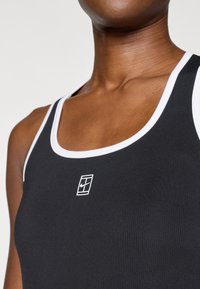 Fekete bordás ujjatlan felső fehér szegéllyel és egy kis fehér logóval a mellkason. Az anyag sima textúrájú és testhez álló kialakítású.