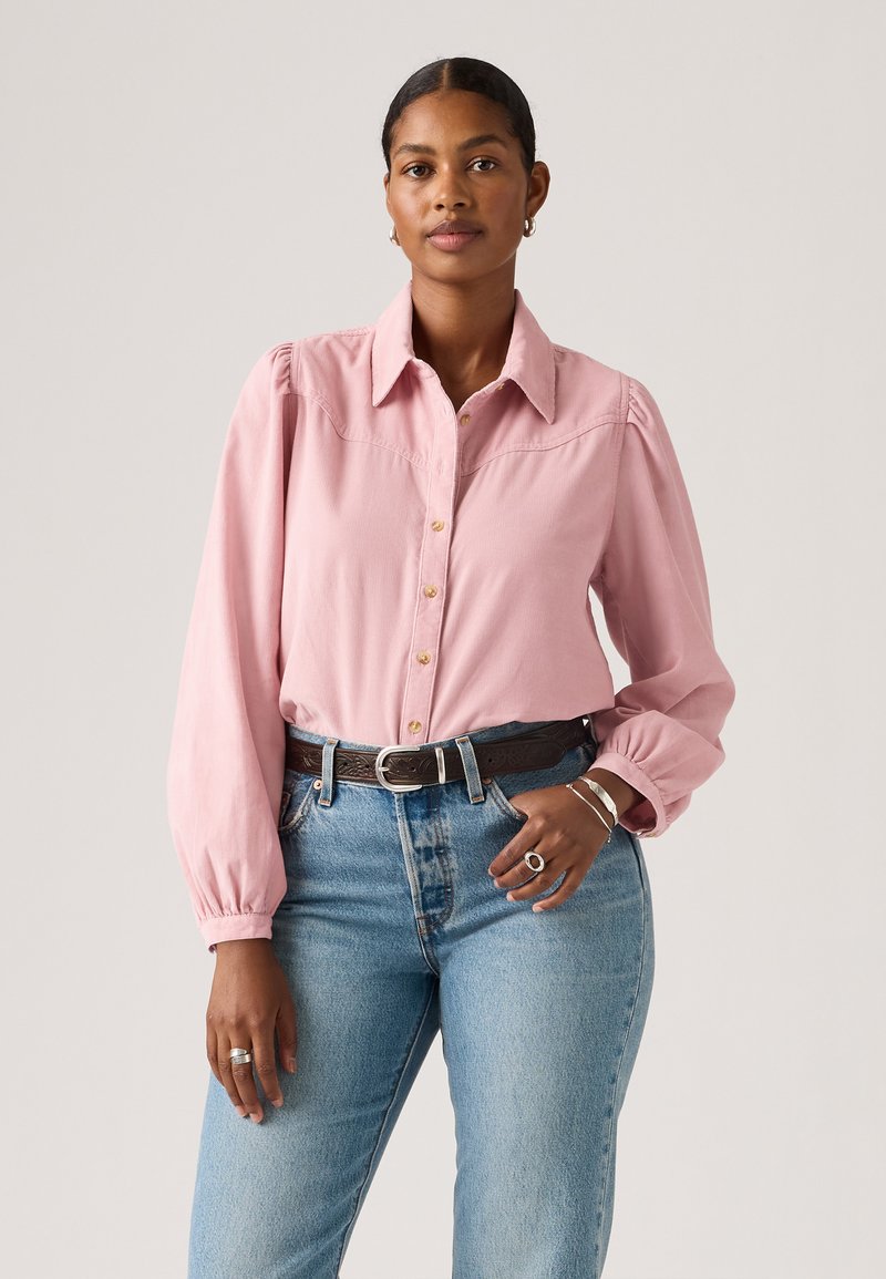 Chemise rose clair à manches longues avec un devant à boutons, des épaules bouffantes et des détails de couture subtils. Associée à un jean bleu taille haute et une ceinture.