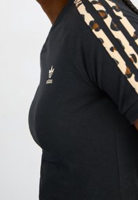 Närbild av person som bär en svart Adidas-tröja med beige och svarta prickade ränder på ärmarna mot en enkel bakgrund.