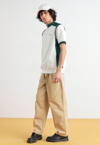 Λευκό και πράσινο αθλητικό πόλο μπλουζάκι με κλείσιμο φερμουάρ, σε συνδυασμό με ανοιχτό μπεζ παντελόνια με φαρδιά γραμμή και μαύρα παπούτσια. Δίδεται έμφαση με ένα ανοιχτό καπέλο.