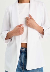 Blazer blanc en tissu léger avec col châle, poignets retroussés et coupe décontractée, associé à un haut blanc et un jean bleu.