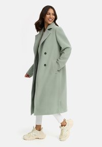 Lichtgroene, dubbelgeknoopte wollen jas met een grote kraag en zijzakken, gedragen over een grijze top en witte broek, gecombineerd met beige sneakers.
