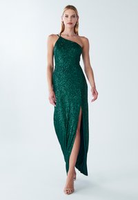 Emeraldgroene sequined avondjurk met een one-shoulder ontwerp, nauwsluitende silhouet en hoge zijsplit. Gladde stof met reflecterende textuur.