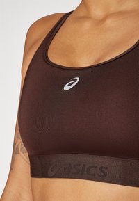 Brązowy stanik sportowy wykonany z gładkiego materiału. Posiada okrągły dekolt, tylną konstrukcję typu racerback oraz szeroki pas z ciemnym logo "asics".