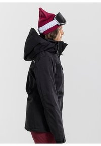 Schwarze Winterjacke mit Kapuze, aus strukturiertem Stoff. Kombiniert mit einer kastanienbraunen Mütze und Skibrille mit weißem Gurt.