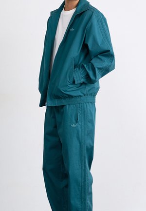 Person iført teal Adidas træningsdragt med jakke lynet halvvejs op og bukser, stående mod en ensfarvet hvid baggrund.